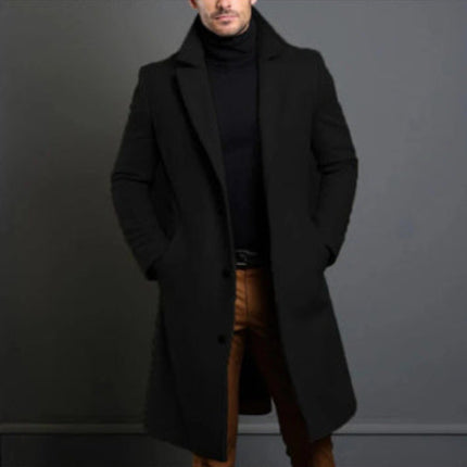 Man wearing a black coat over a black turtleneck and brown pants against a gray background