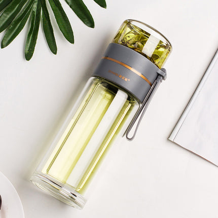 Clear glass water bottle with gray lid and handle on a white surface with green leaves.