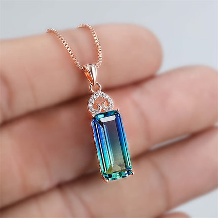 Blue gemstone pendant on a rose gold chain held in a hand against a neutral background