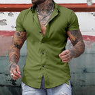 Man wearing a green button-up shirt with tattoos visible, standing against a rustic wall.