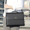 Person holding a black leather briefcase with a brand logo, blurred background