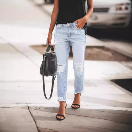 Person wearing light blue ripped jeans and black sandals, holding a black handbag on a blurred street background.