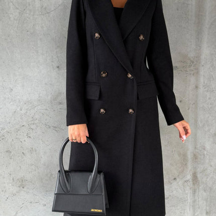 Person wearing a black coat holding a black handbag against a gray background