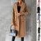 Woman wearing a long beige coat holding a black handbag against a gray background.