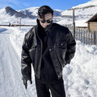 Person wearing a black leather jacket and sunglasses standing in a snowy landscape with mountains in the background.