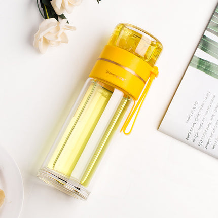 Yellow and clear glass water bottle on a light surface with flowers and a book in the background